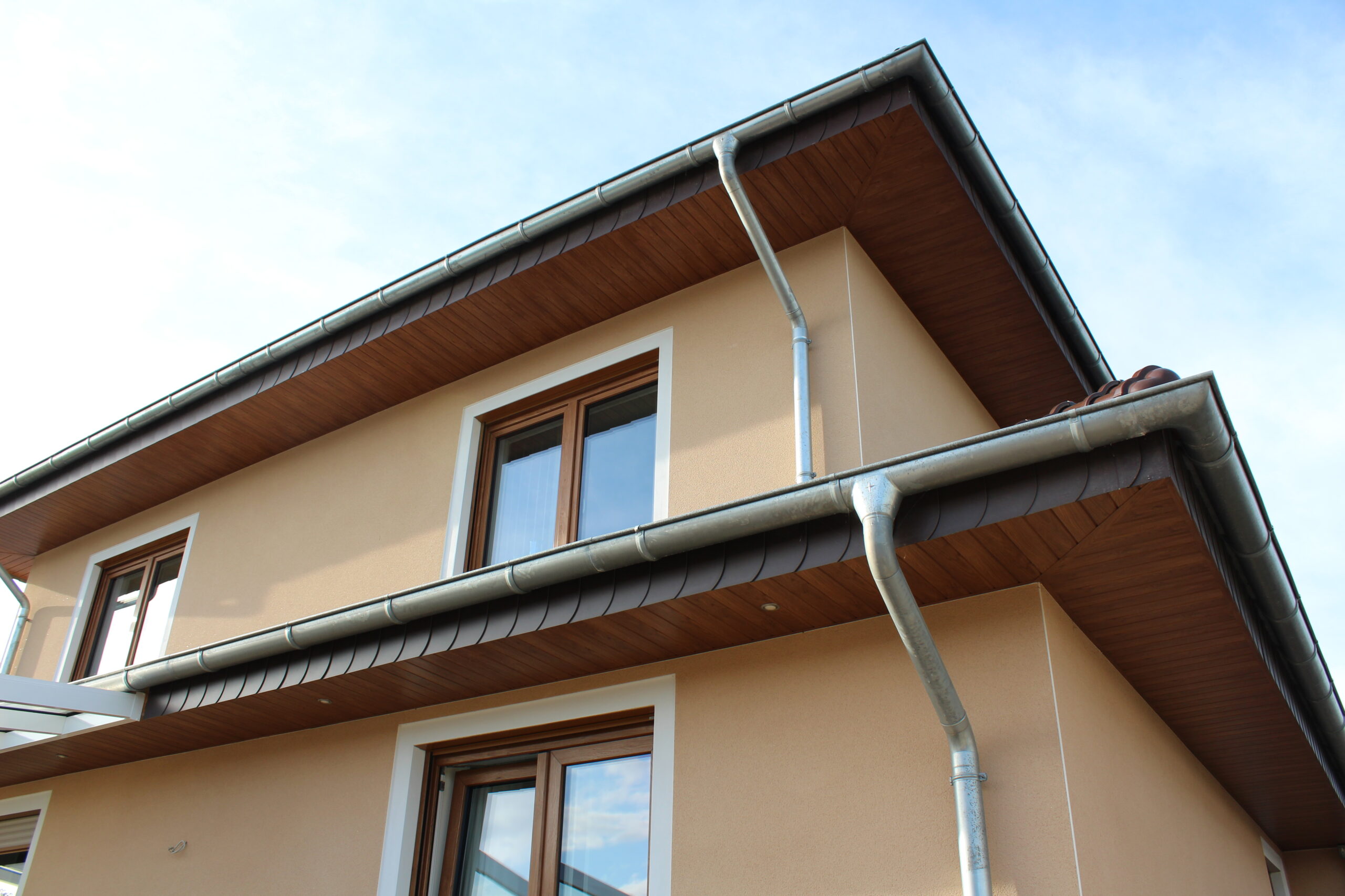 Ein modernes Wohnhaus mit beigen Wänden, braunen Fensterrahmen und einem grauen Dach mit Regenrinnen aus Metall.