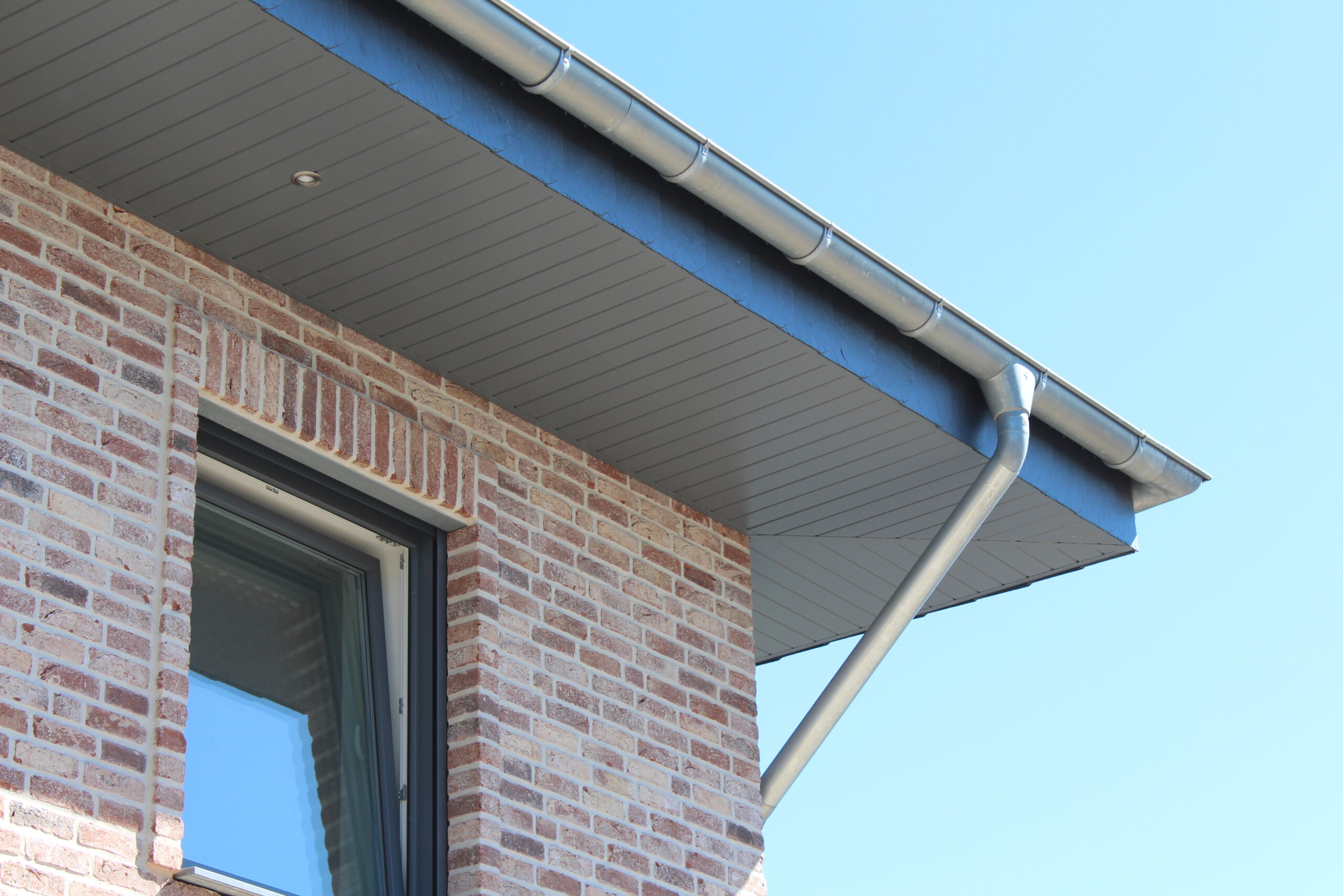 Ein modernes Ziegelhaus mit grauem Dachüberstand und Dachrinne, sowie einem Fenster im oberen Stockwerk, an einem sonnigen Tag.