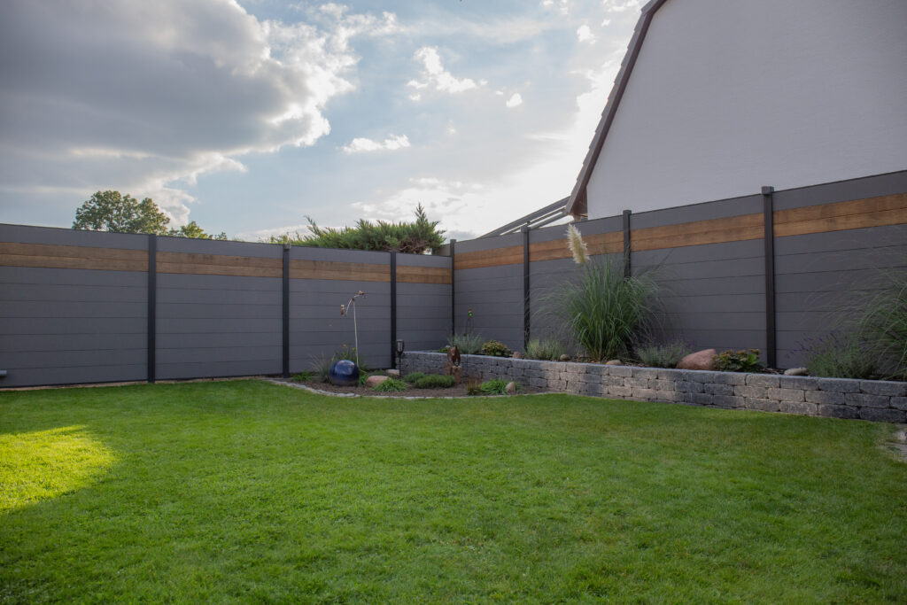 Ein moderner Garten mit einem grauen und hölzernem Sichtschutzzaun, einer gepflegten Rasenfläche und einem kleinen bepflanzten Blumenbeet mit dekorativen Elementen und Natursteinen, im Hintergrund ein weißes Hausdach sichtbar.
