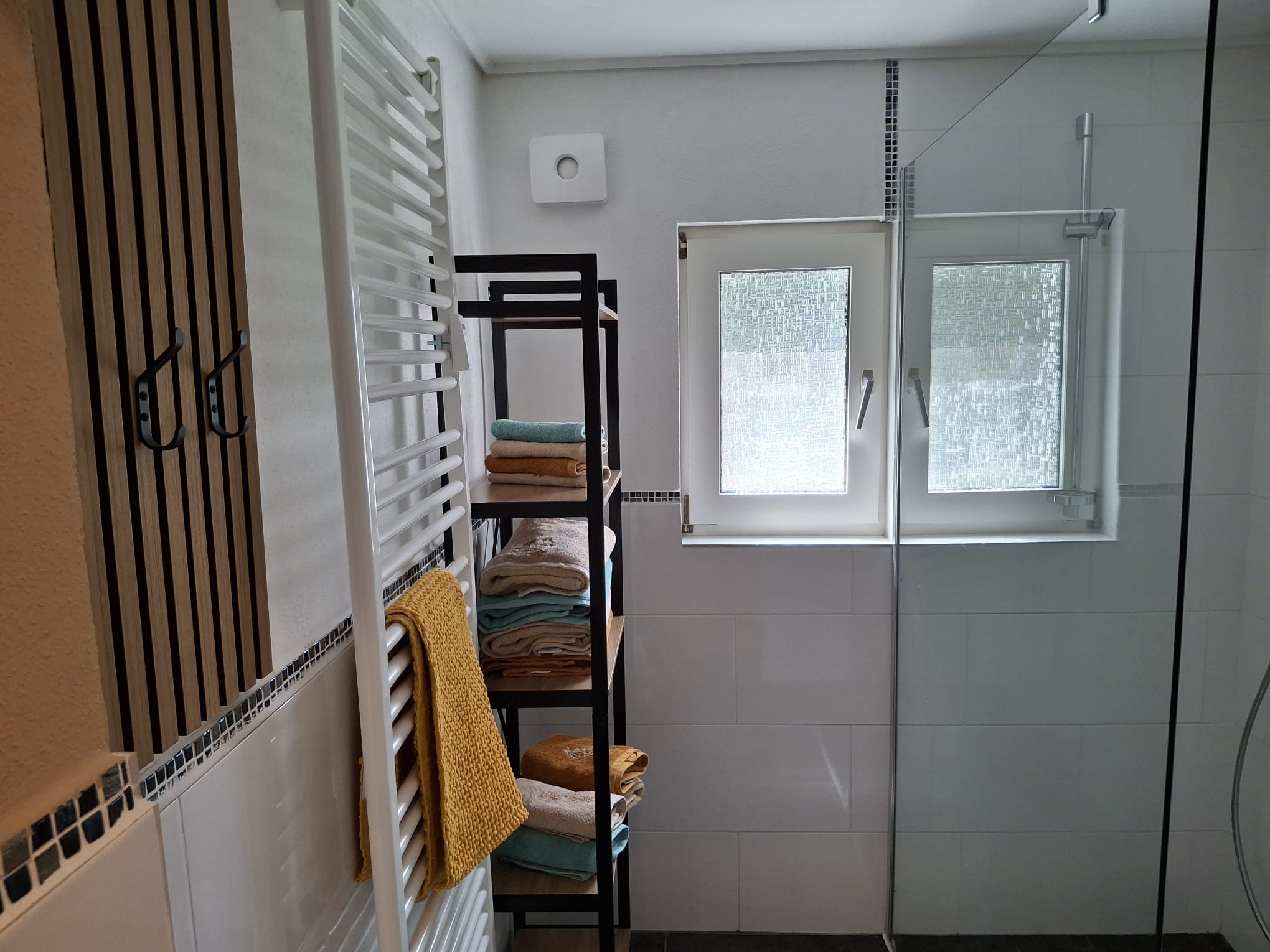 Modernes Badezimmer mit einem Handtuchheizkörper, einem Regal mit gestapelten Handtüchern und einem Fenster mit Milchglas, das teilweise von einer Duschwand verdeckt wird.