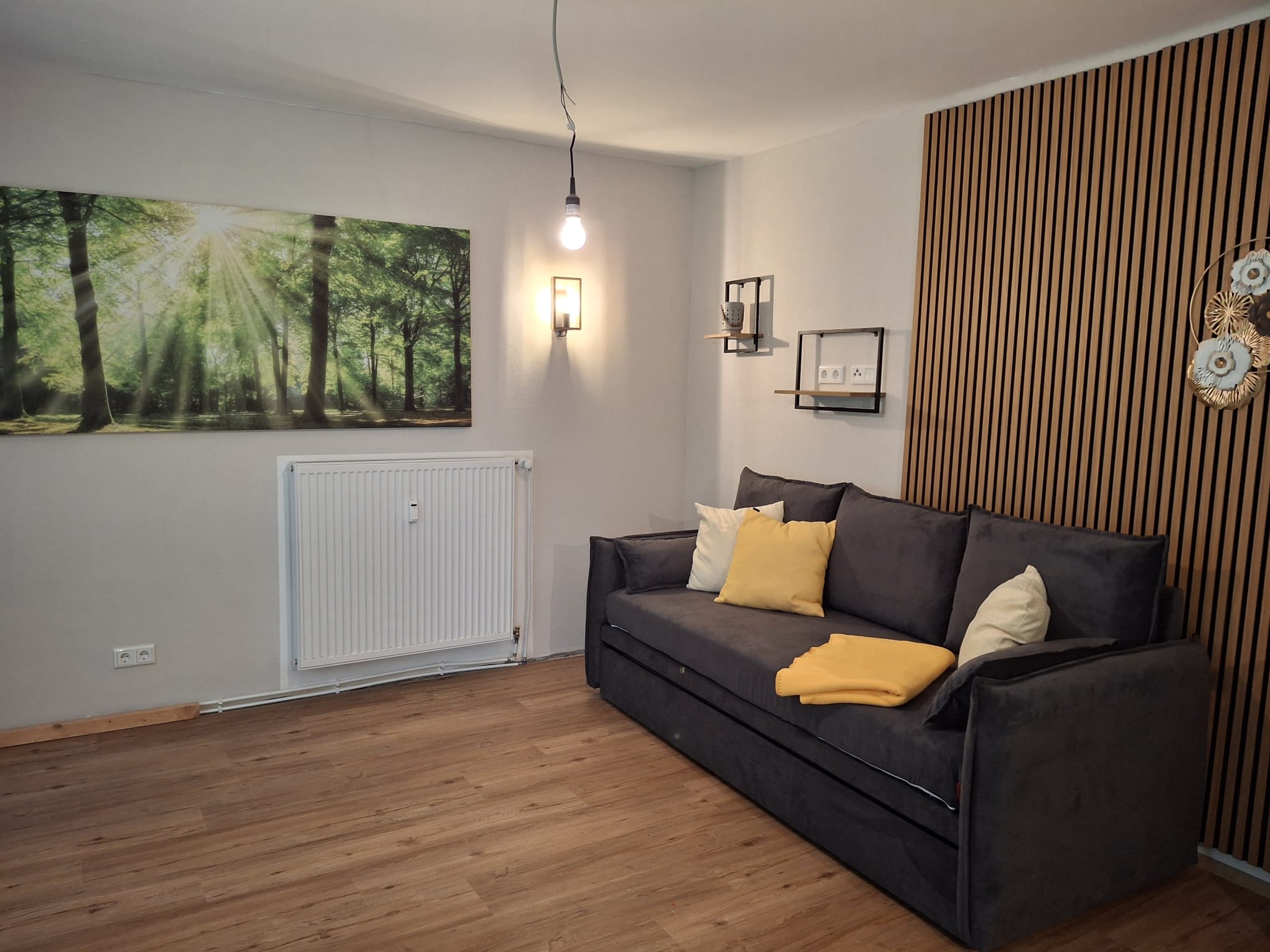 Ein modern eingerichtetes Wohnzimmer mit Holzfußboden, einem dunkelgrauen Sofa und gelben Kissen. An der Wand hängt ein großes Bild von einem Wald mit Sonnenstrahlen. Einfache Wandlampen und eine minimalistische Regalstruktur zieren die weiße Wand. Ein vertikales Holzgitter verleiht dem Raum eine stilvolle Note.