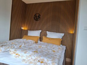 Ein modernes Schlafzimmer mit zwei Einzelbetten, die weiße und gelbe florale Bettwäsche sowie gelbe Kissen haben, vor einer Holzwand mit vertikalen Streifen und zwei Wandleuchten.