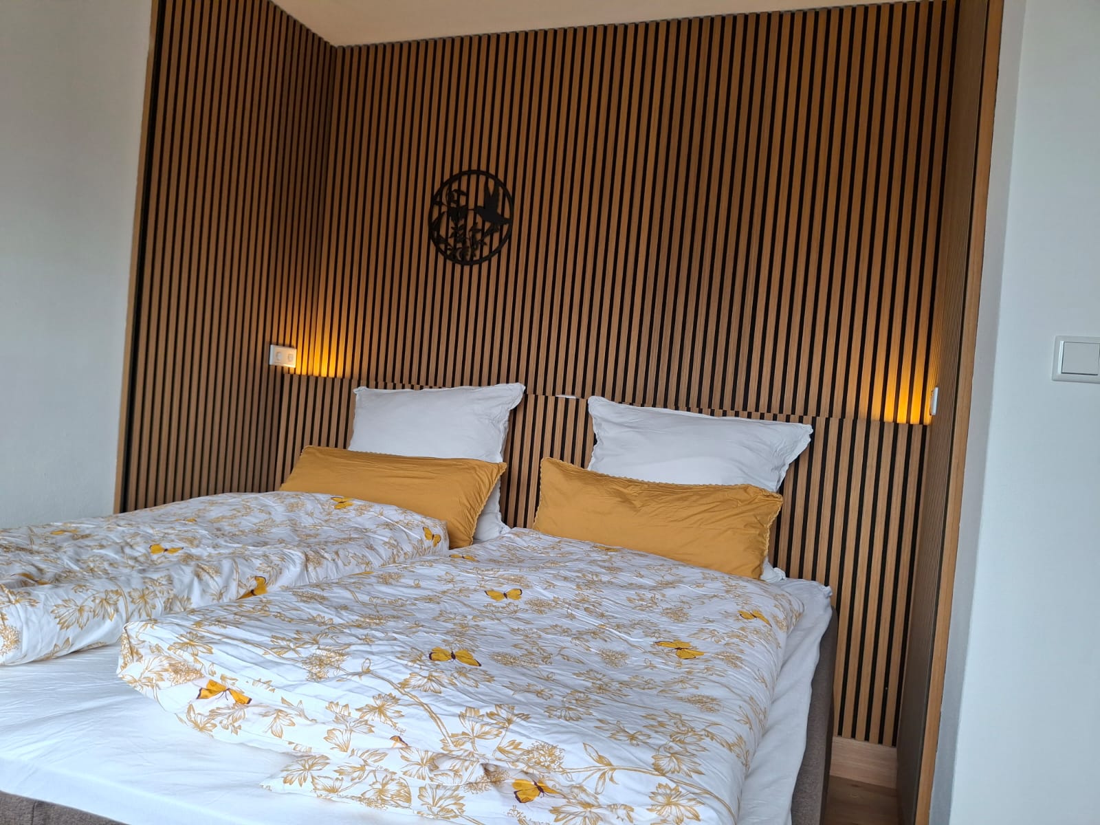 Ein modernes Schlafzimmer mit zwei Einzelbetten, die weiße und gelbe florale Bettwäsche sowie gelbe Kissen haben, vor einer Holzwand mit vertikalen Streifen und zwei Wandleuchten.