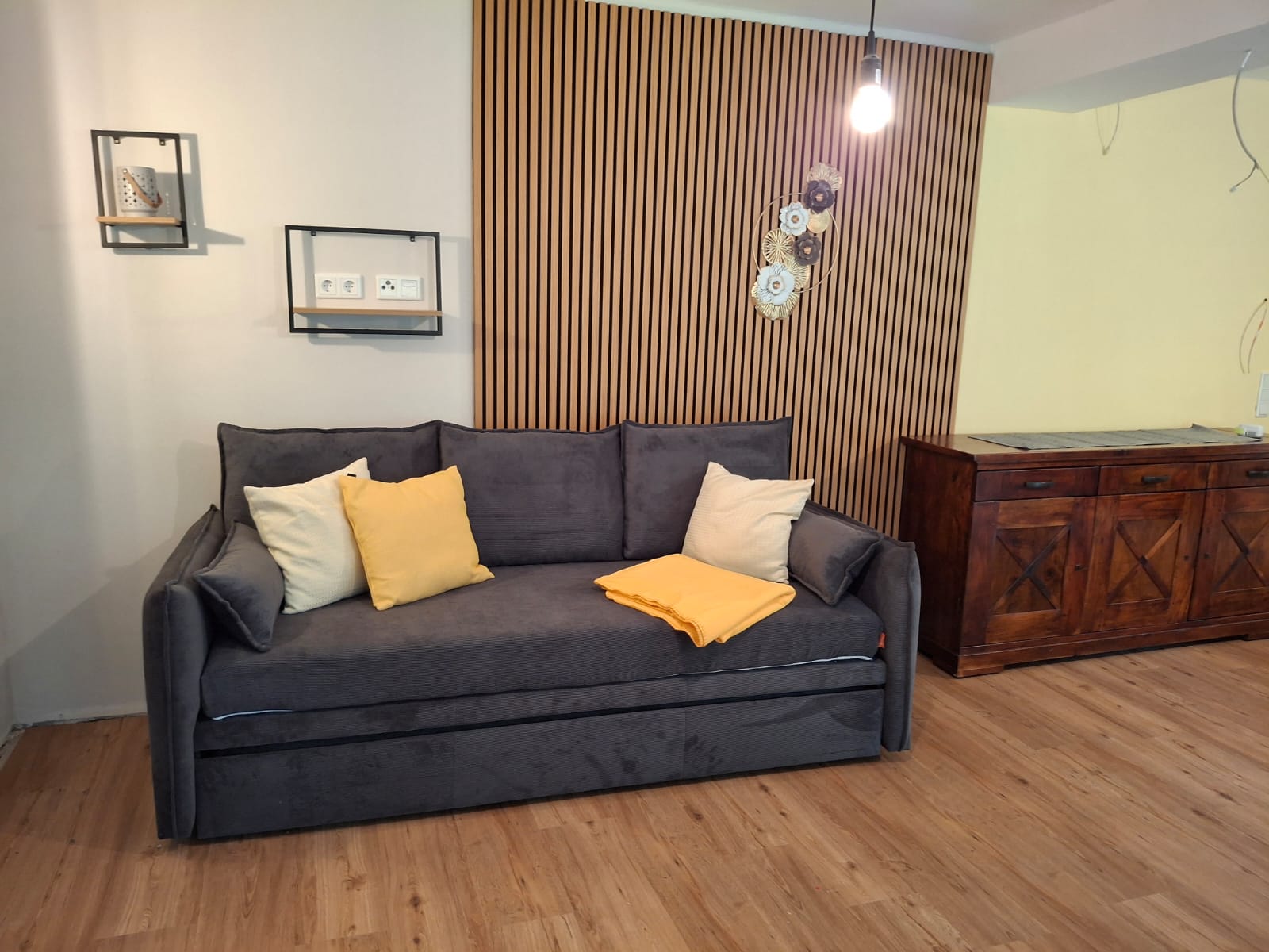 Modernes Wohnzimmer mit einem grauen Sofa, dekoriert mit gelben und weißen Kissen. An der Wand befindet sich eine Holzvertäfelung und daneben steht eine dunkle Kommode. Eine Lampe hängt von der Decke und zwei Regale sind an der weißen Wand befestigt.