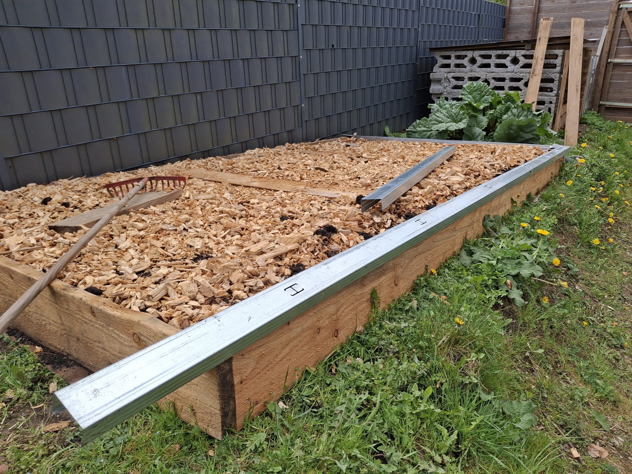 Ein Hochbeet gefüllt mit Holzspänen, darüber liegt eine Harke. Daneben befinden sich Metallleisten und einige Pflanzen, darunter Löwenzahn, die um das Beet herum wachsen. Im Hintergrund sind Metall- und Holzwände sowie eine kleine Kompost- oder Steinbox zu sehen.