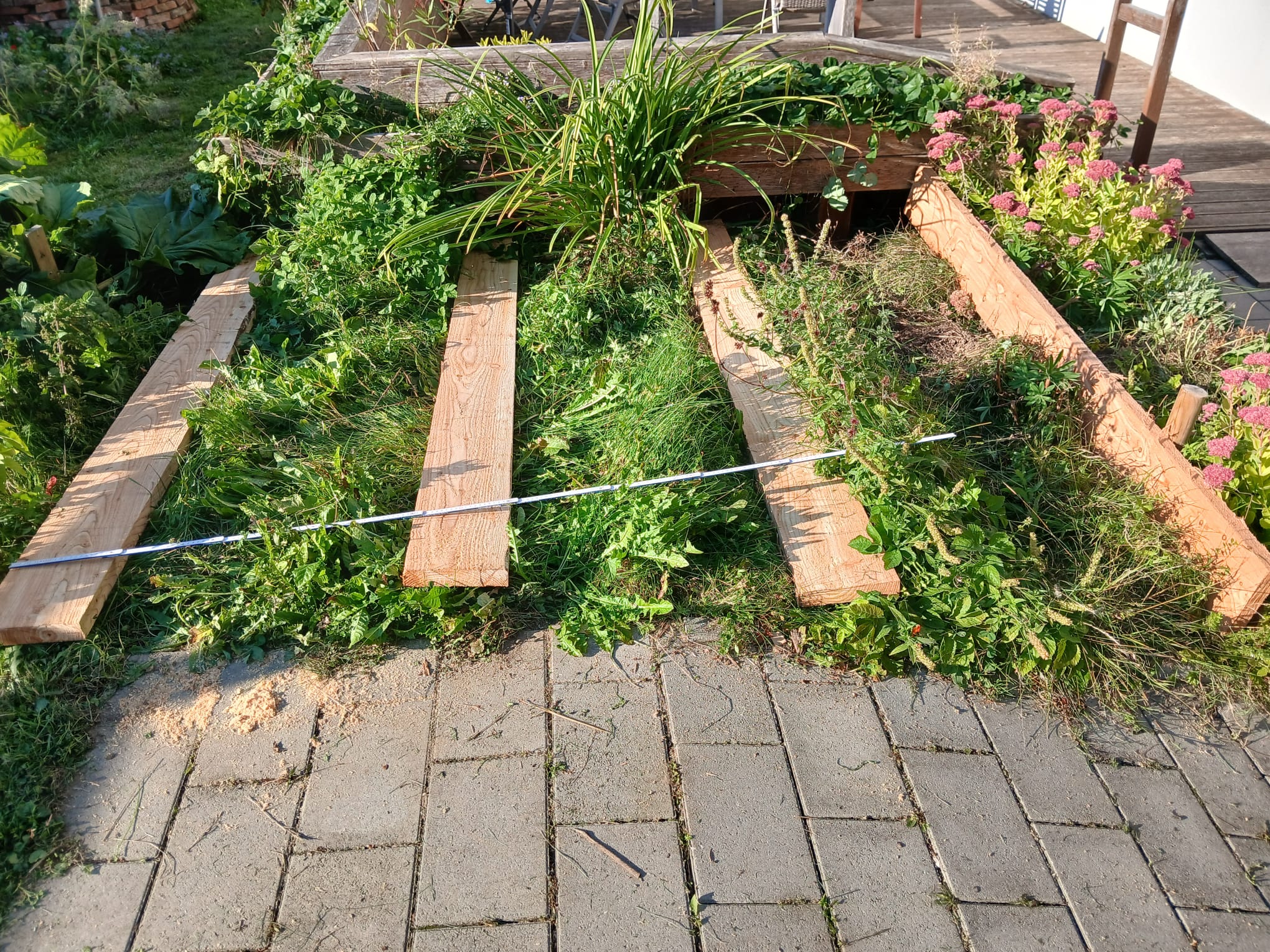 Holzbretter liegen horizontal auf einem mit Gras und Unkraut bedeckten Boden vor einer erhöhten Blumenbeeteinfassung, daneben ein mit Blumen bewachsener Streifen und ein Metalllineal.