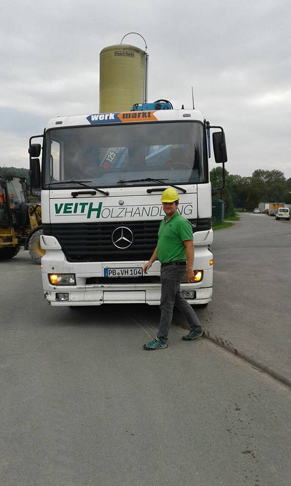 Ein Mann mit gelbem Bauhelm steht vor einem weißen Lastwagen mit der Aufschrift 'Veith Holzhandlung'.