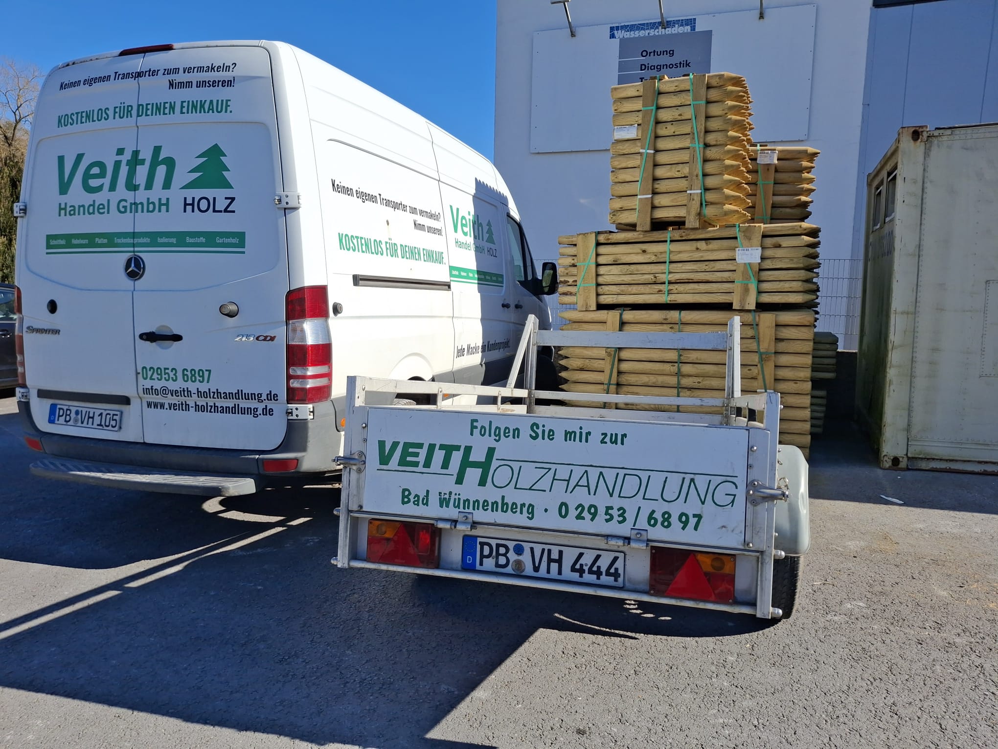 Ein weißer Lieferwagen der Firma Veith Holzhandlung parkt neben einem Anhänger mit gestapelten Holzbalken. Der Anhänger trägt ebenfalls die Aufschrift der Firma. Im Hintergrund ist ein Lagergebäude zu sehen.