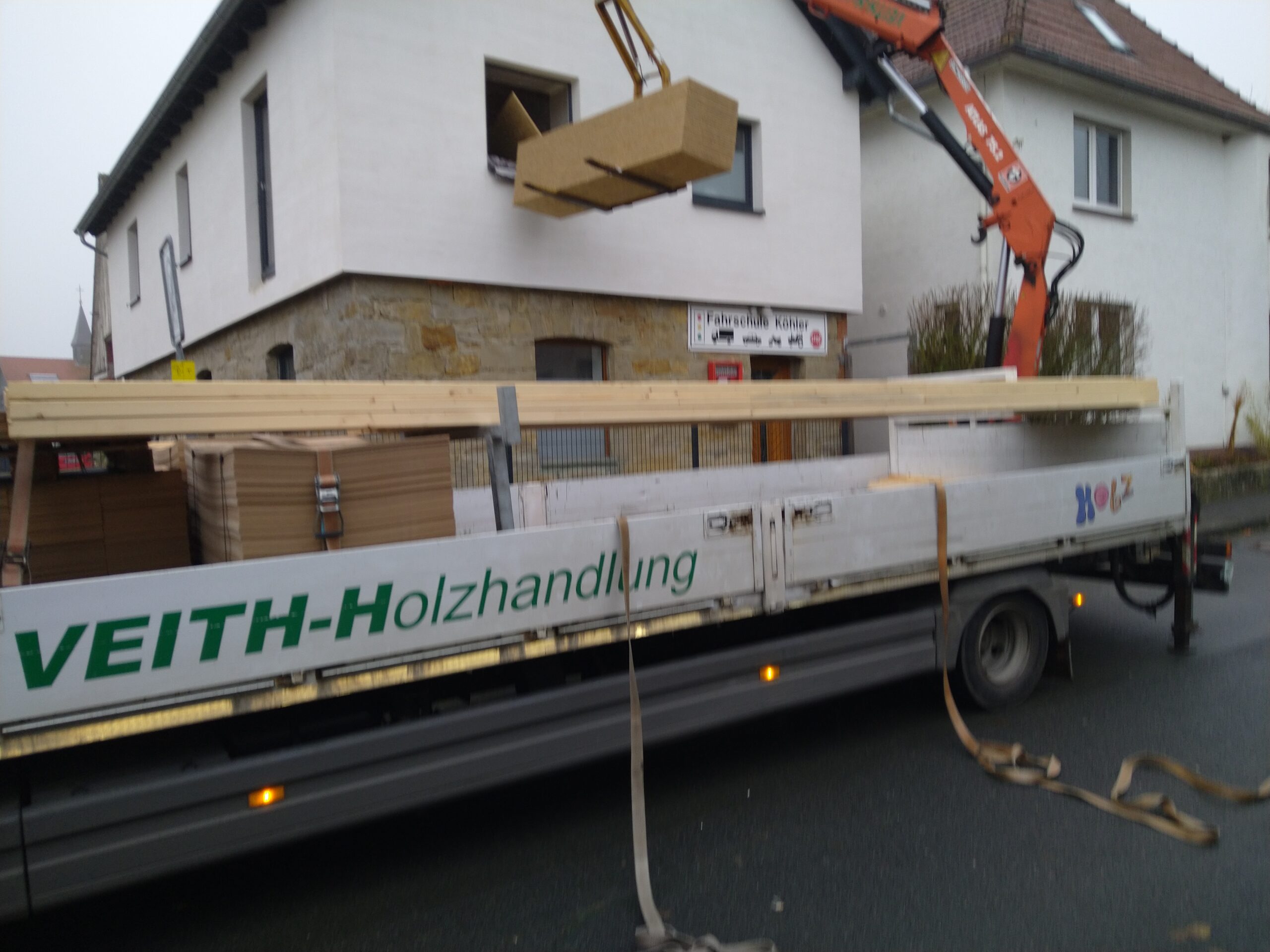 Ein Lastwagen von Veith-Holzhandlung mit Holz beladen wird vor einem Gebäude mit Hilfe eines Krans entladen.