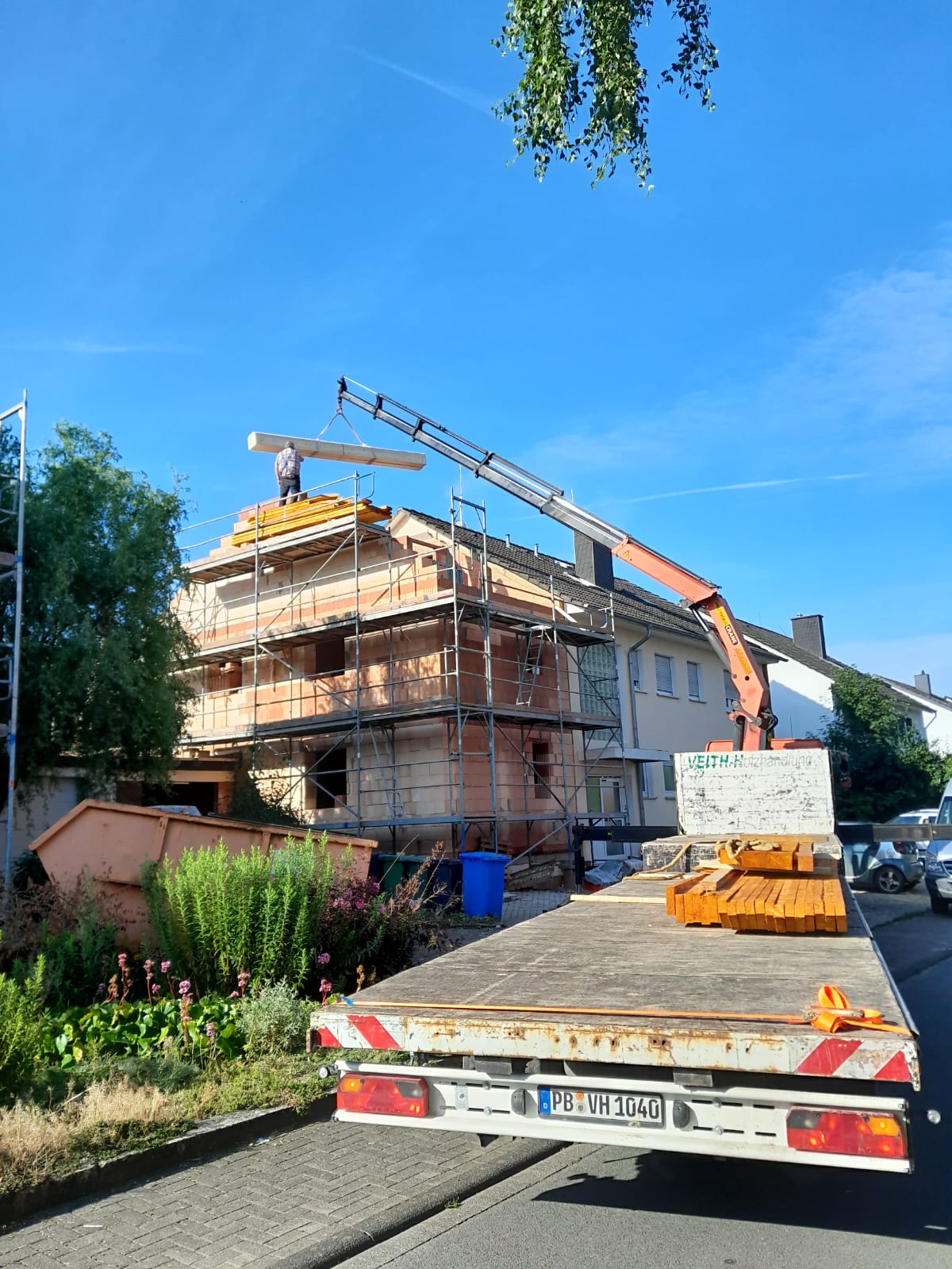 Ein Baustellen-Lkw steht vor einem im Bau befindlichen Haus, das mit einem Gerüst umgeben ist. Ein Kran auf dem Lkw hebt einen großen Holzbalken auf das Dach des Hauses. Ein Arbeiter ist auf dem Dach sichtbar. Der Himmel ist klar und blau.