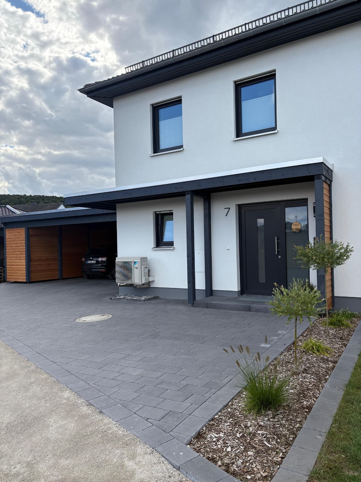 Modernes zweistöckiges Haus mit weißer Fassade und grauem Dach, davor ein gepflasterter Weg und ein kleiner Gartenbereich mit Pflanzen, sowie ein überdachter Carport links im Bild.