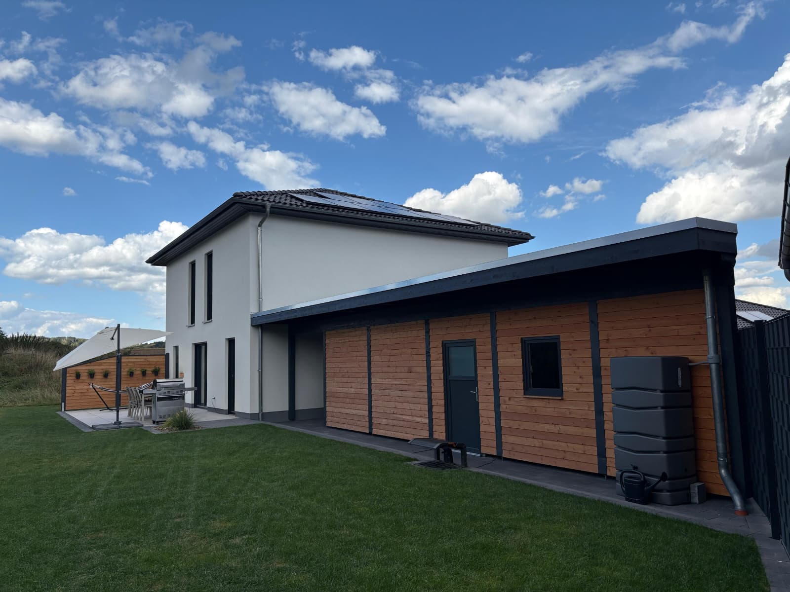 Modernes zweistöckiges Haus mit einer Fassade in Weiß und Holz, flankiert von einem flachen Anbau, umgeben von einem gepflegten Rasen unter einem blauen Himmel mit Wolken.