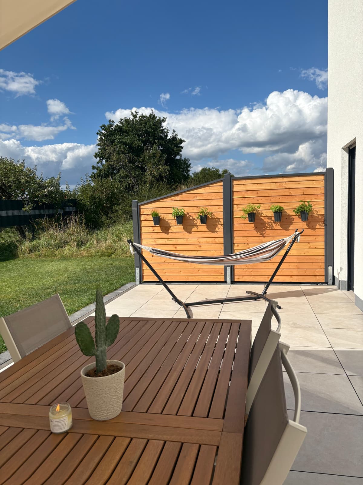Eine Terrasse mit Holztisch, auf dem ein Kaktus und eine Kerze stehen, daneben zwei Stühle, ein leeres Hängemattengestell und ein Holzsichtschutz mit Pflanztöpfen, hinter dem sich ein Garten mit Bäumen und blauem Himmel befindet.