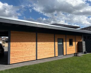 Holzverkleidetes Nebengebäude mit flachem Dach, grauer Tür und kleinem Fenster, daneben stehen Regentonnen; seitlich ist ein Auto unter einem Carport geparkt, auf dem Rasen im Vordergrund ist sonniges Wetter mit Wolken am Himmel zu sehen.