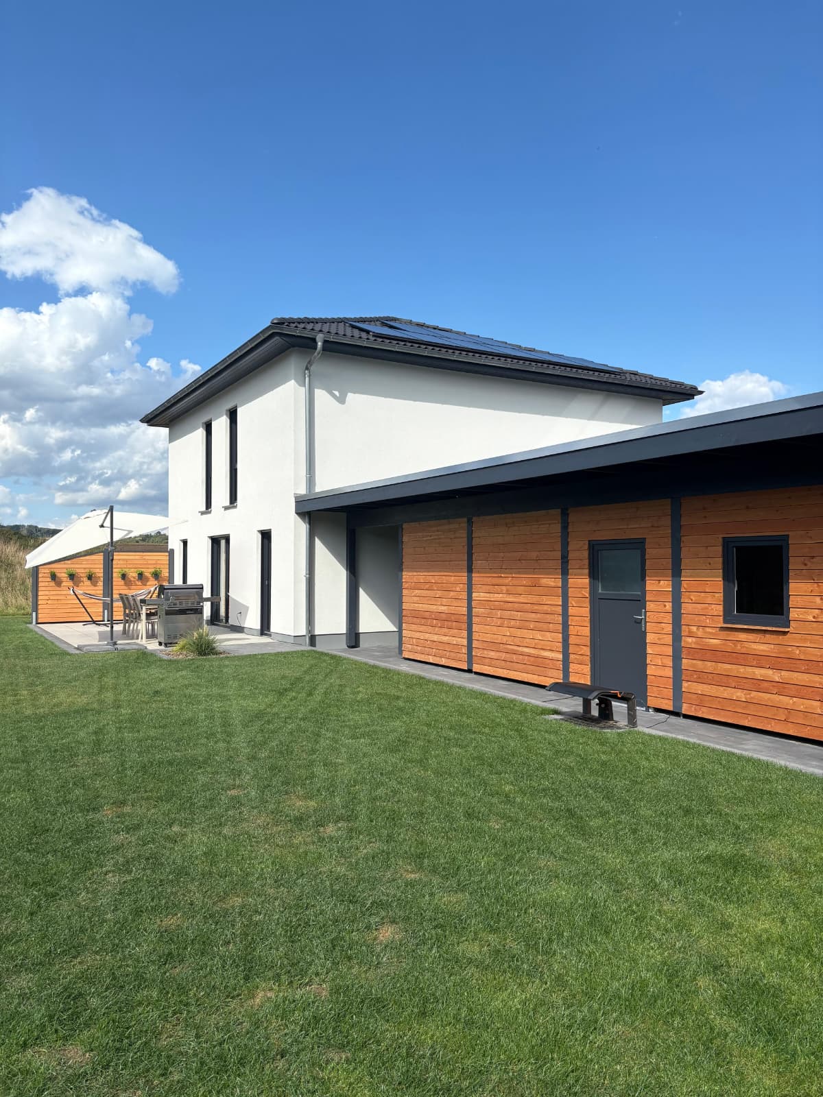 Modernes zweistöckiges Gebäude mit weißer Fassade, schwarzen Fenstern und einem flachen Dach, daneben ein Anbau mit Holzverkleidung, auf einer gepflegten Rasenfläche unter blauem Himmel mit Wolken.