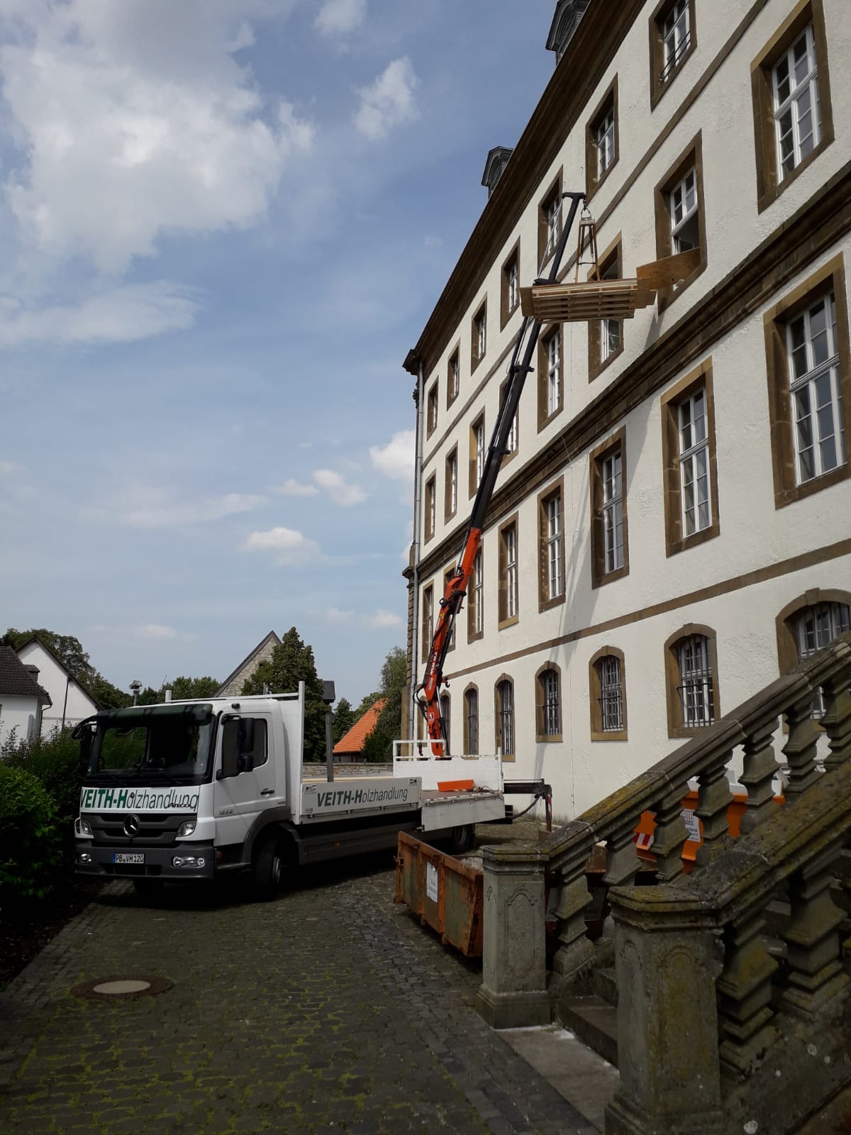Ein LKW der Firma Veith Holzhandlung steht vor einem mehrstöckigen Gebäude und hebt mit einem Kran Paletten in ein Fenster im oberen Stockwerk.