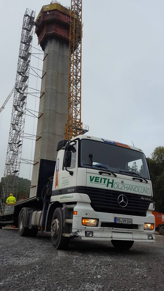 Ein Lastwagen der Firma Veith Holzhandlung steht auf einer Baustelle, im Hintergrund ist ein hoher Betonturm mit Gerüst und Kran zu sehen.