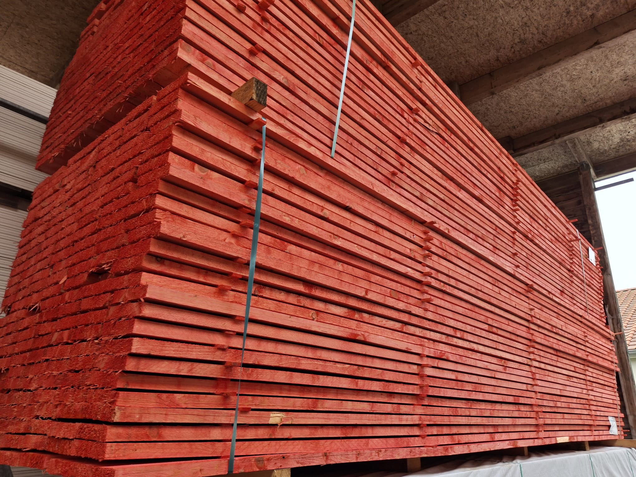Ein großer Stapel roter Holzbretter in einer Lagerhalle, ordentlich gestapelt und mit einem blauen Band gesichert.