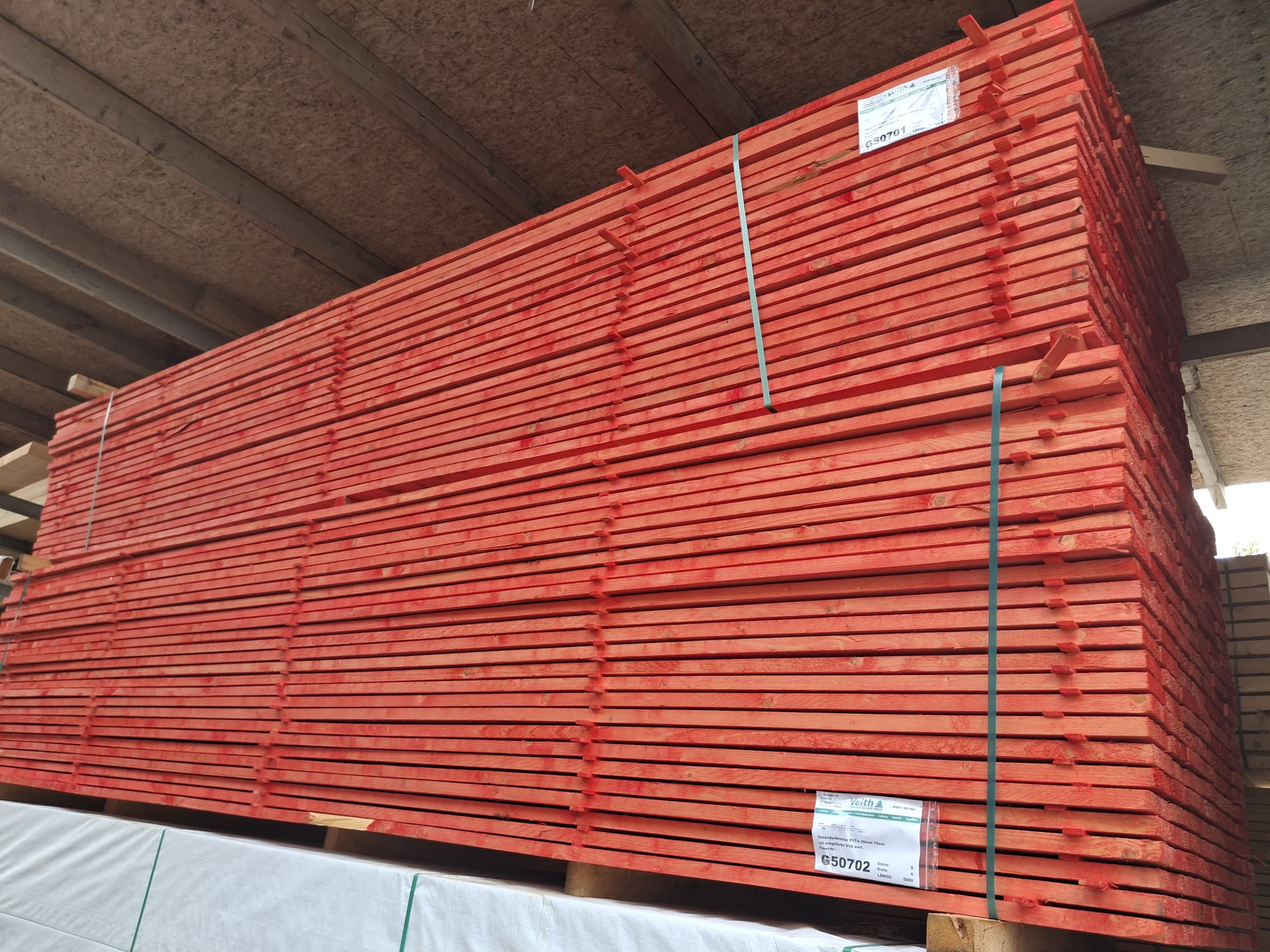 Ein großer Stapel roter Holzlatten, ordentlich gestapelt und gebündelt, steht in einem Lagerraum mit sichtbarer Dachkonstruktion aus Holz.
