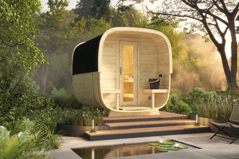 Kleine, freistehende Sauna aus Holz mit Panoramafenster und Terrasse, umgeben von Bäumen und Garten, am Rande eines Teichs.