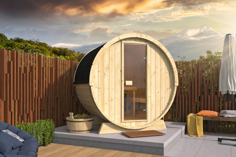 Fassförmige Sauna auf einer Terrasse, umgeben von Holzzaun und Gartenmöbeln, Berglandschaft im Hintergrund.