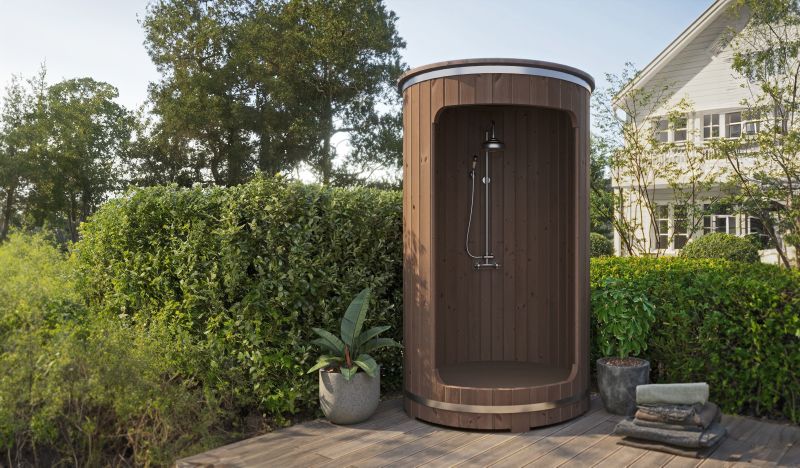 Runde Außendusche aus Holz auf einer Terrasse, umgeben von Pflanzen und dichtem Strauchwerk.