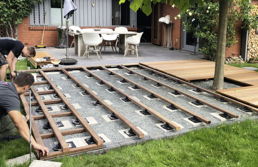 Zwei Personen bauen eine Holzterrasse im Garten, mit einer Konstruktion aus Holzbalken über Kiesboden.