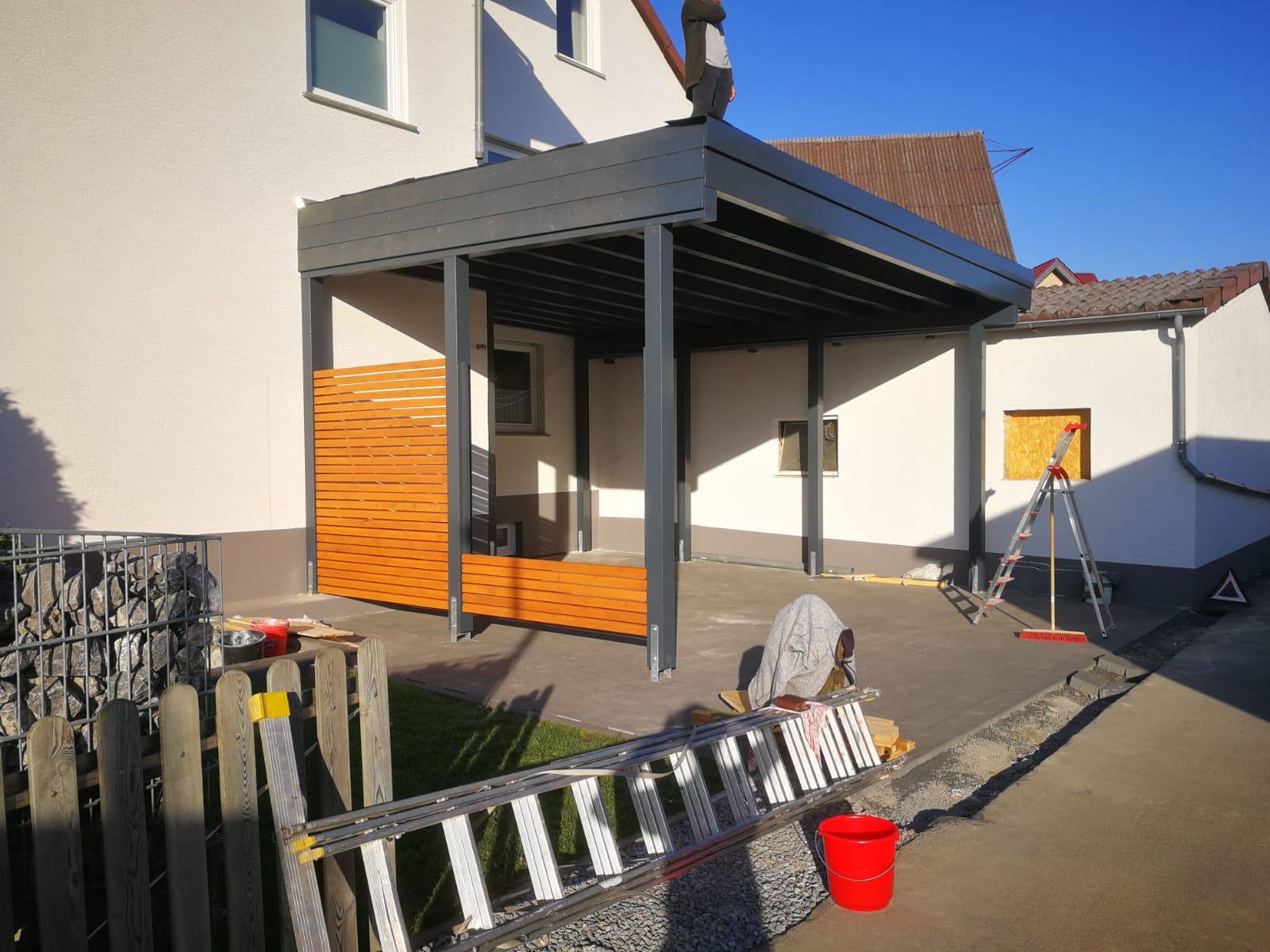 Eine überdachte Terrasse mit Holzelementen, Metallstützen und Bauwerkzeugen wie einer Leiter und einem Eimer im Vordergrund.