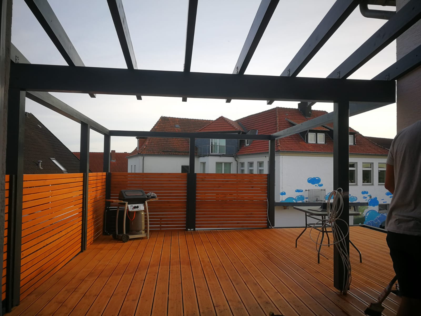 Holzterrasse mit Pergola, Holzzaun und Grill, im Hintergrund Häuser mit roten Dächern.