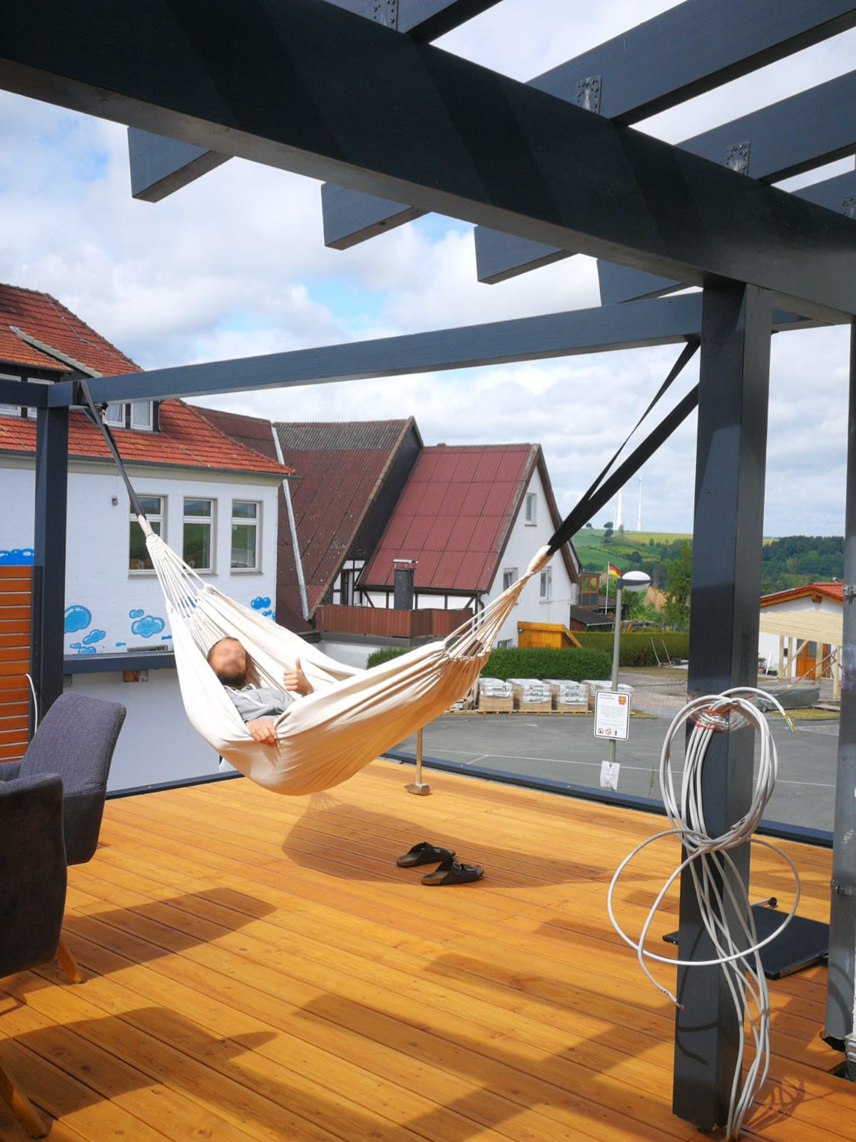 Person in einer Hängematte auf einer Holzterrasse, umgeben von Häusern und blauem Himmel.