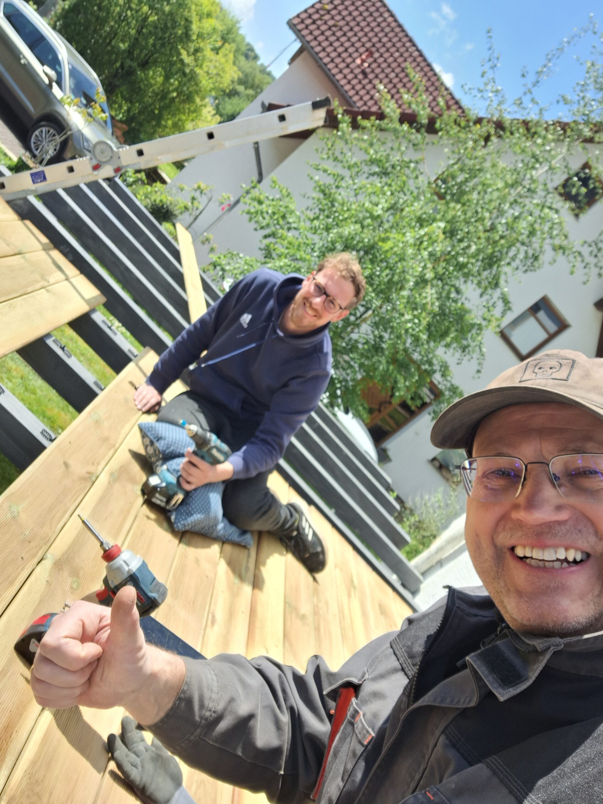 Zwei Männer arbeiten lächelnd auf einer Holzterrasse; der Mann im Vordergrund zeigt Daumen hoch.