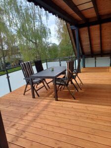 Ein Holzbalkon mit einem rechteckigen Tisch und sechs Gartenstühlen, umgeben von Glasgeländern.