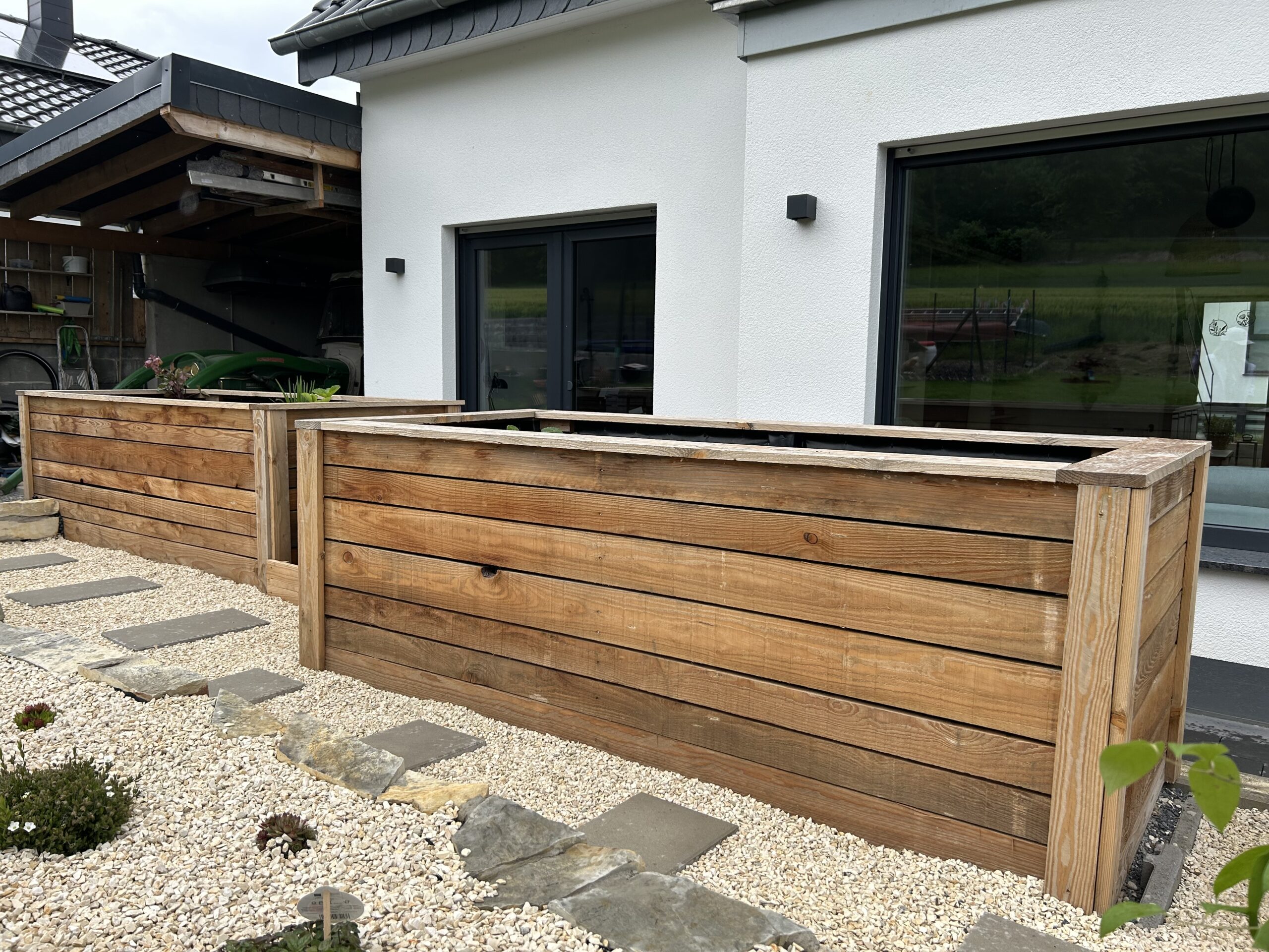 Holzhochbeete auf einer Terrasse neben einem modernen weißen Haus, umgeben von Kieselsteinen und einigen Pflanzen.