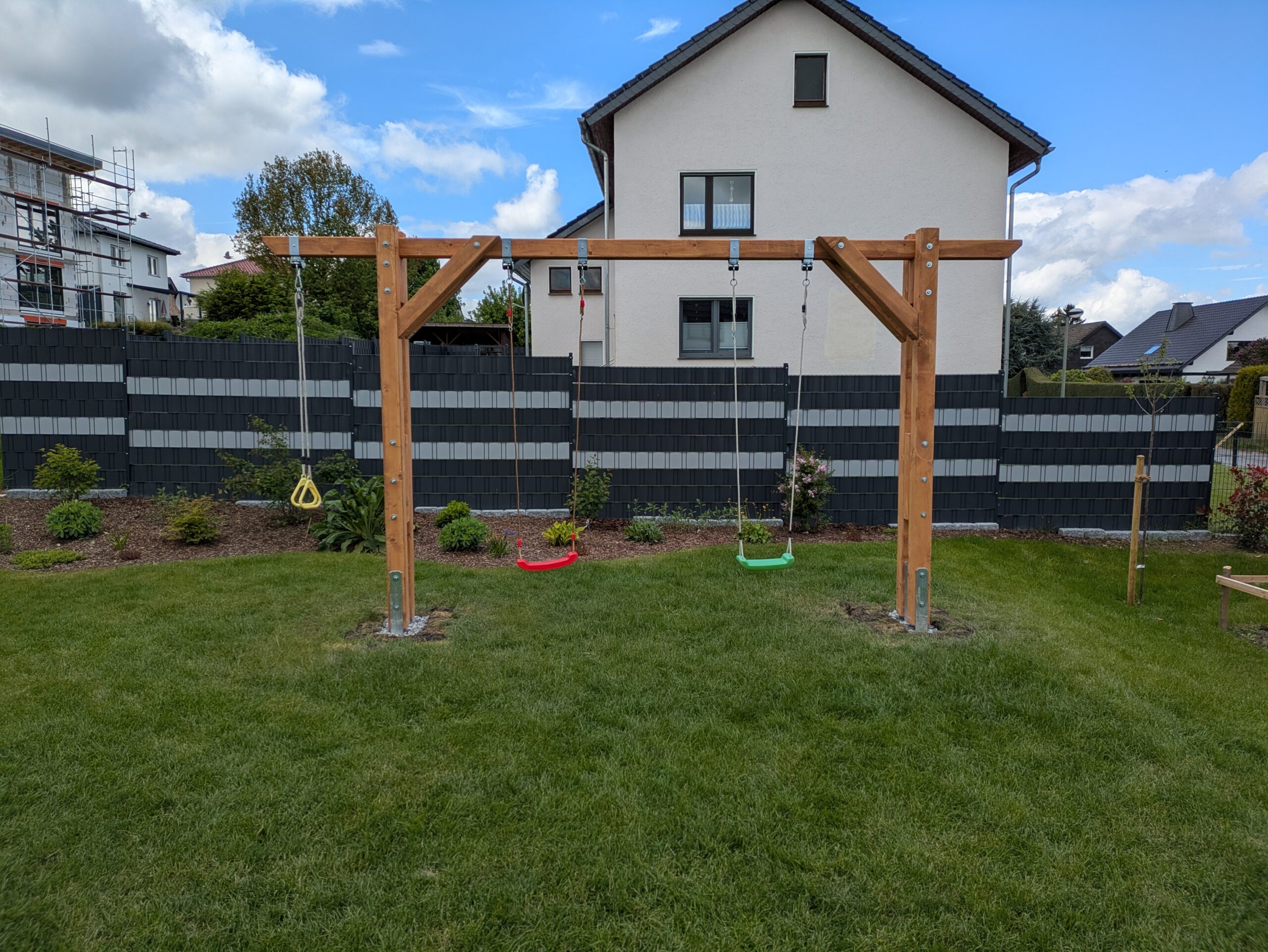 Holzspielgerüst mit zwei Schaukeln und Ringen auf einem Rasen vor einem Wohnhaus.