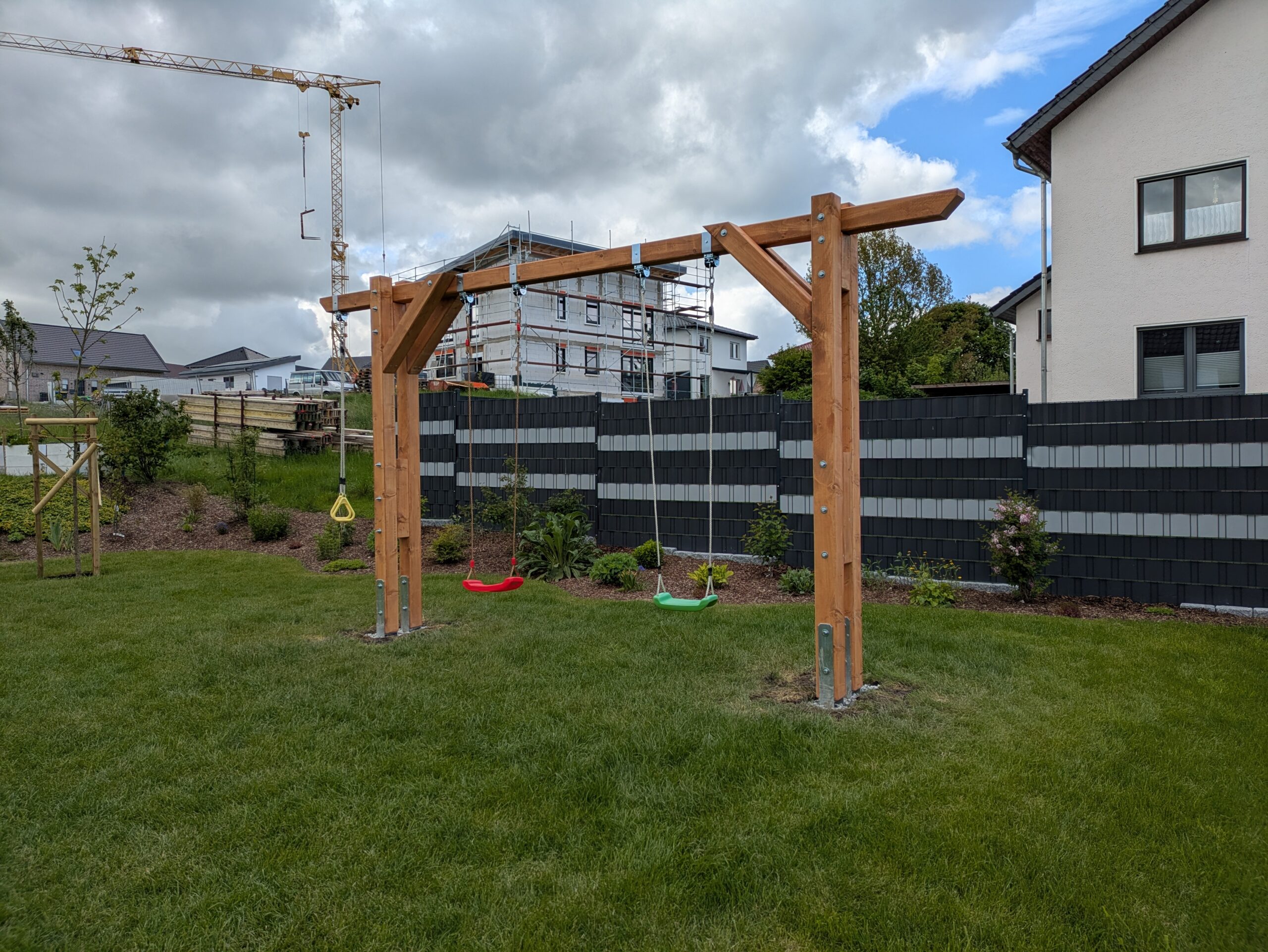 Holz-Schaukelgestell mit zwei Schaukeln auf einer Rasenfläche, umgeben von Häusern und einem Baukran im Hintergrund.