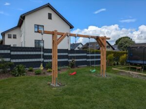 Holzschaukel mit drei Schaukeln auf einer Rasenfläche, im Hintergrund ein weißes Haus und ein Zaun.