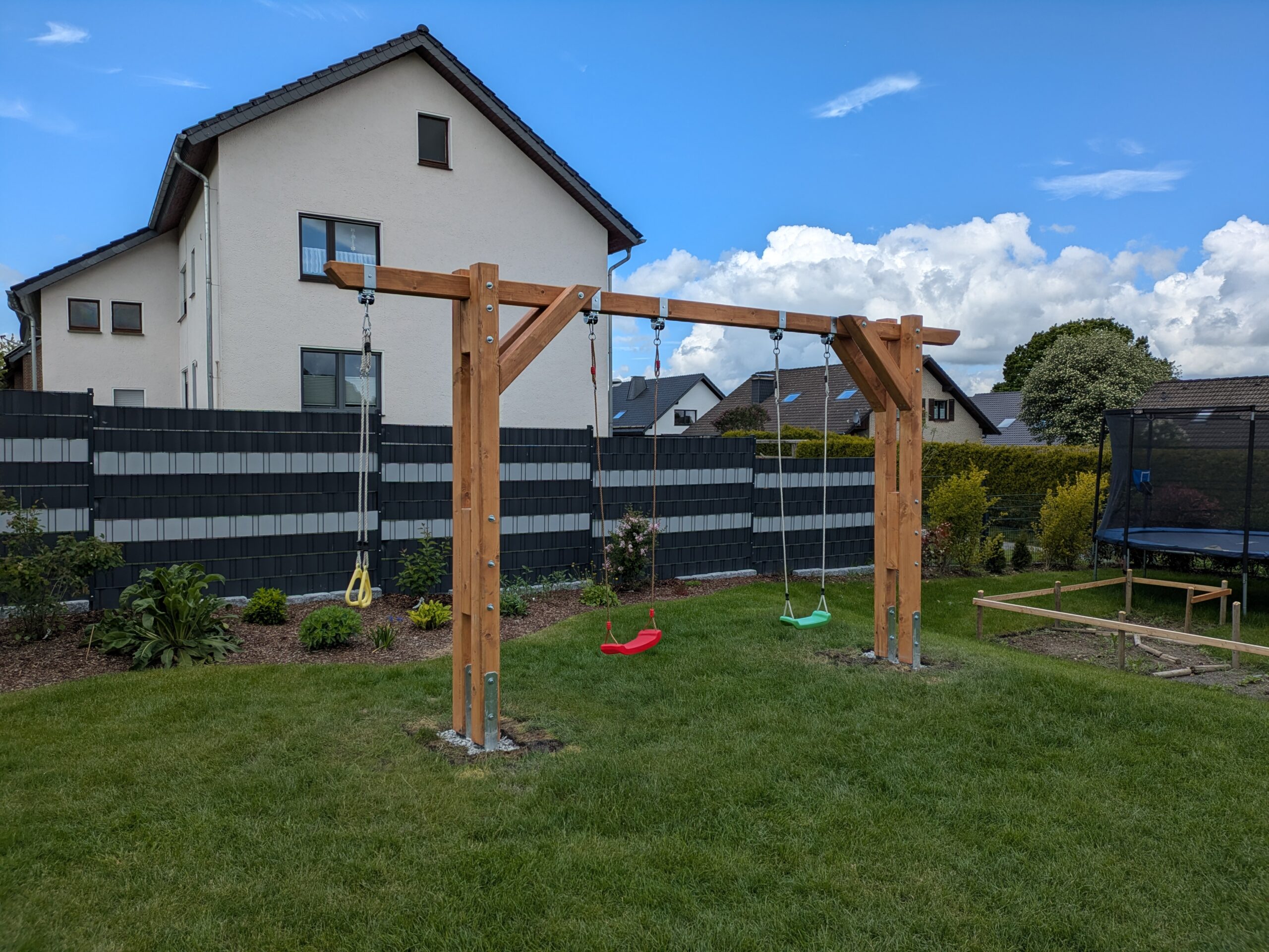 Holzschaukel mit drei Schaukeln auf einer Rasenfläche, im Hintergrund ein weißes Haus und ein Zaun.