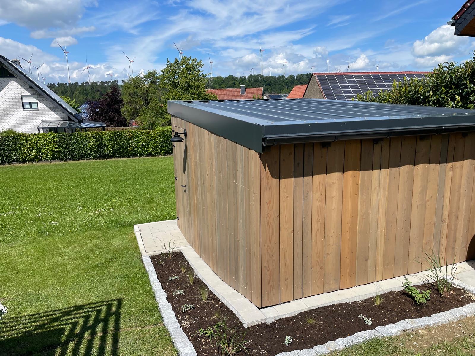 Holzschuppen im Garten mit Solarpanelen auf dem Dach, umgeben von einer Wiese und gepflasterten Wegen.