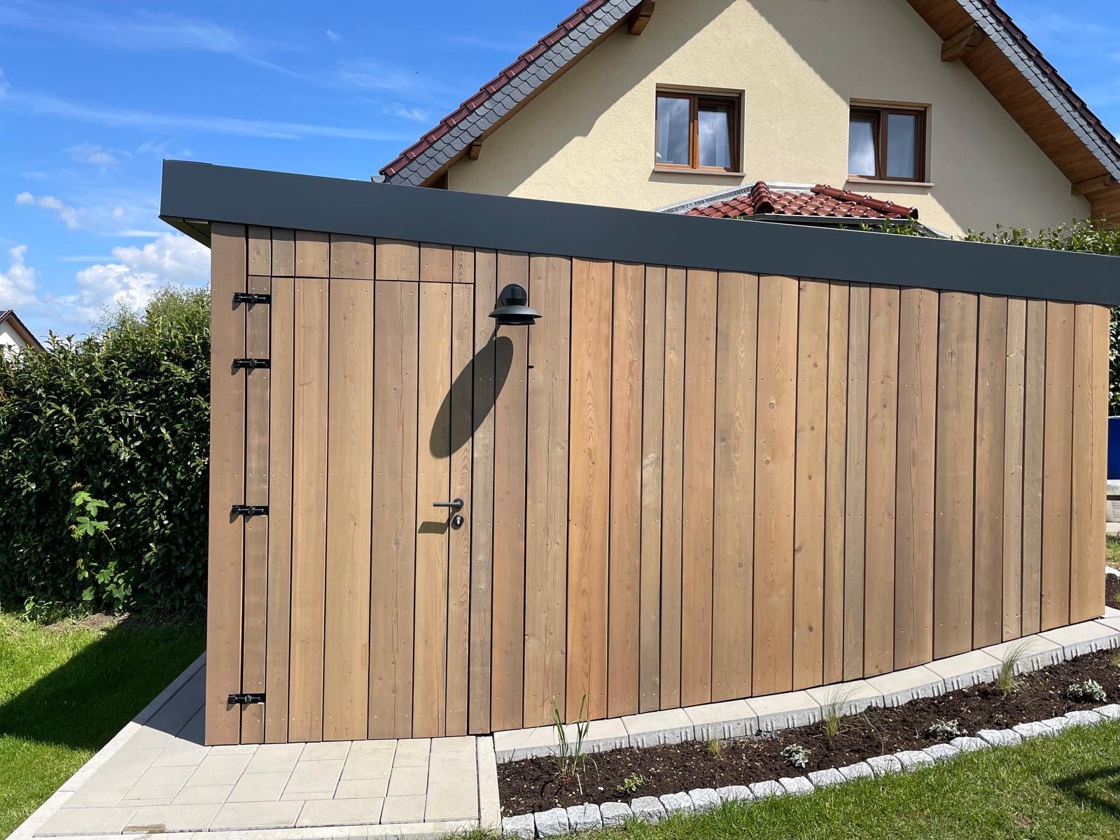 Holzschuppen mit geneigtem Dach und schwarzer Lampe, im Garten eines Hauses mit beigem Putz und roten Dachziegeln.