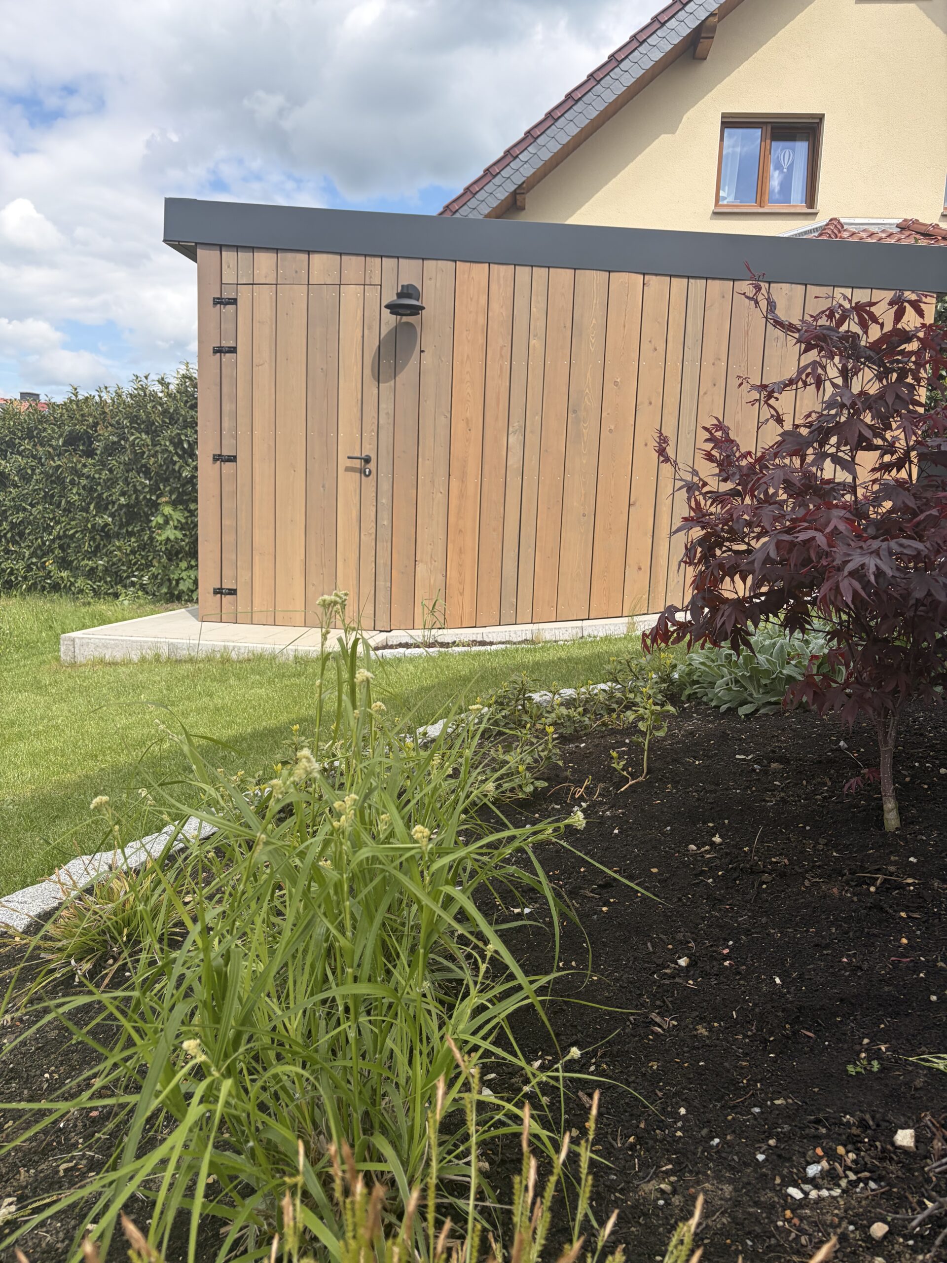 Holzschuppen im Garten mit angrenzendem Beet und grünem Rasen, im Hintergrund ein Haus mit Giebeldach.