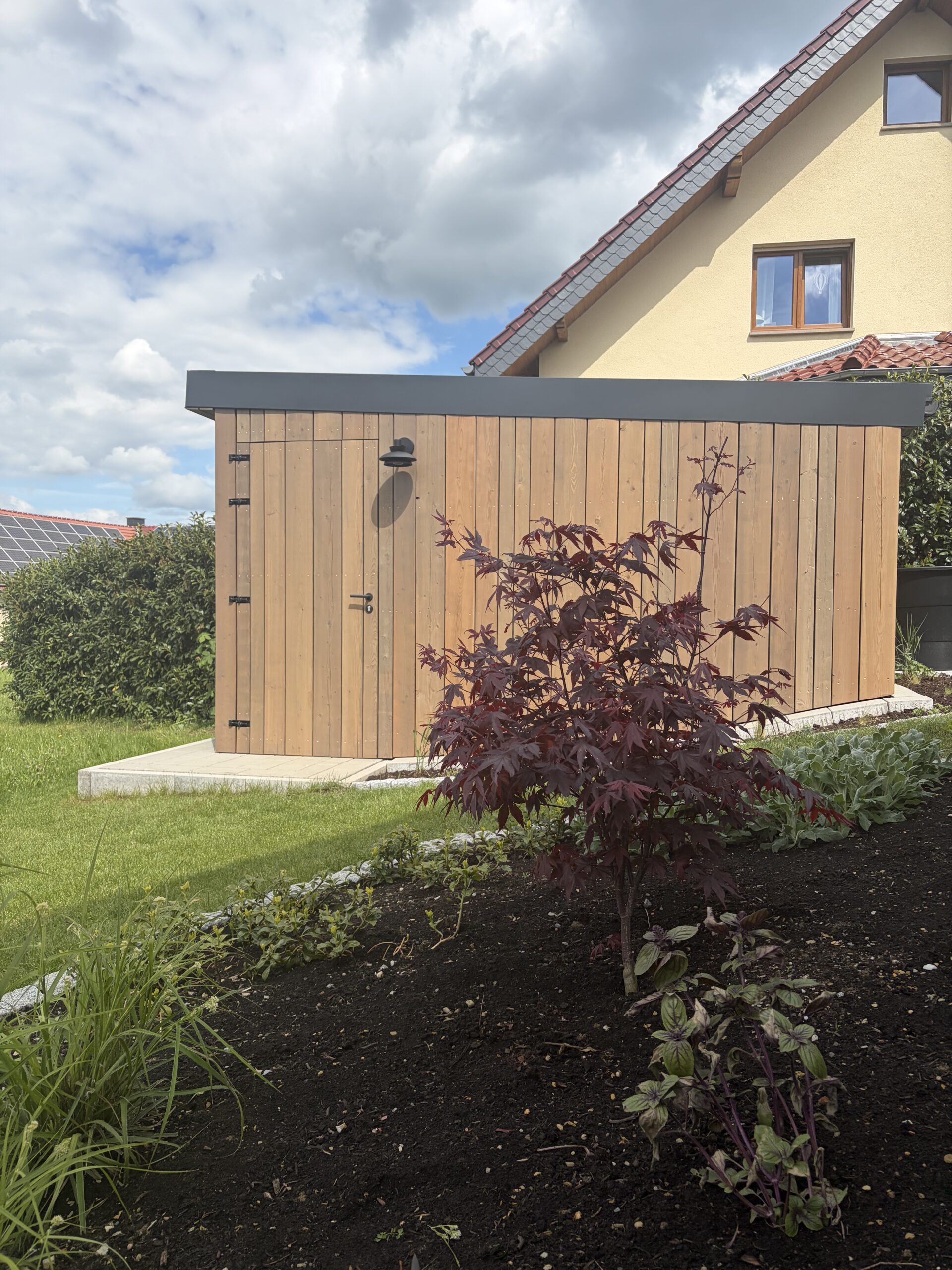 Holzgartenhaus mit Flachdach in einem Garten, umgeben von blühenden Pflanzen und einem Haus im Hintergrund.