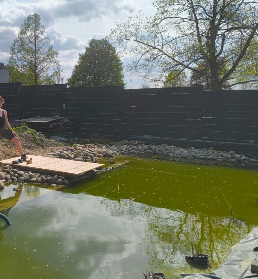 Eine Person sitzt auf einem Holzsteg am Rand eines kleinen Teichs in einem Garten mit grünem Wasser und Bäumen im Hintergrund.