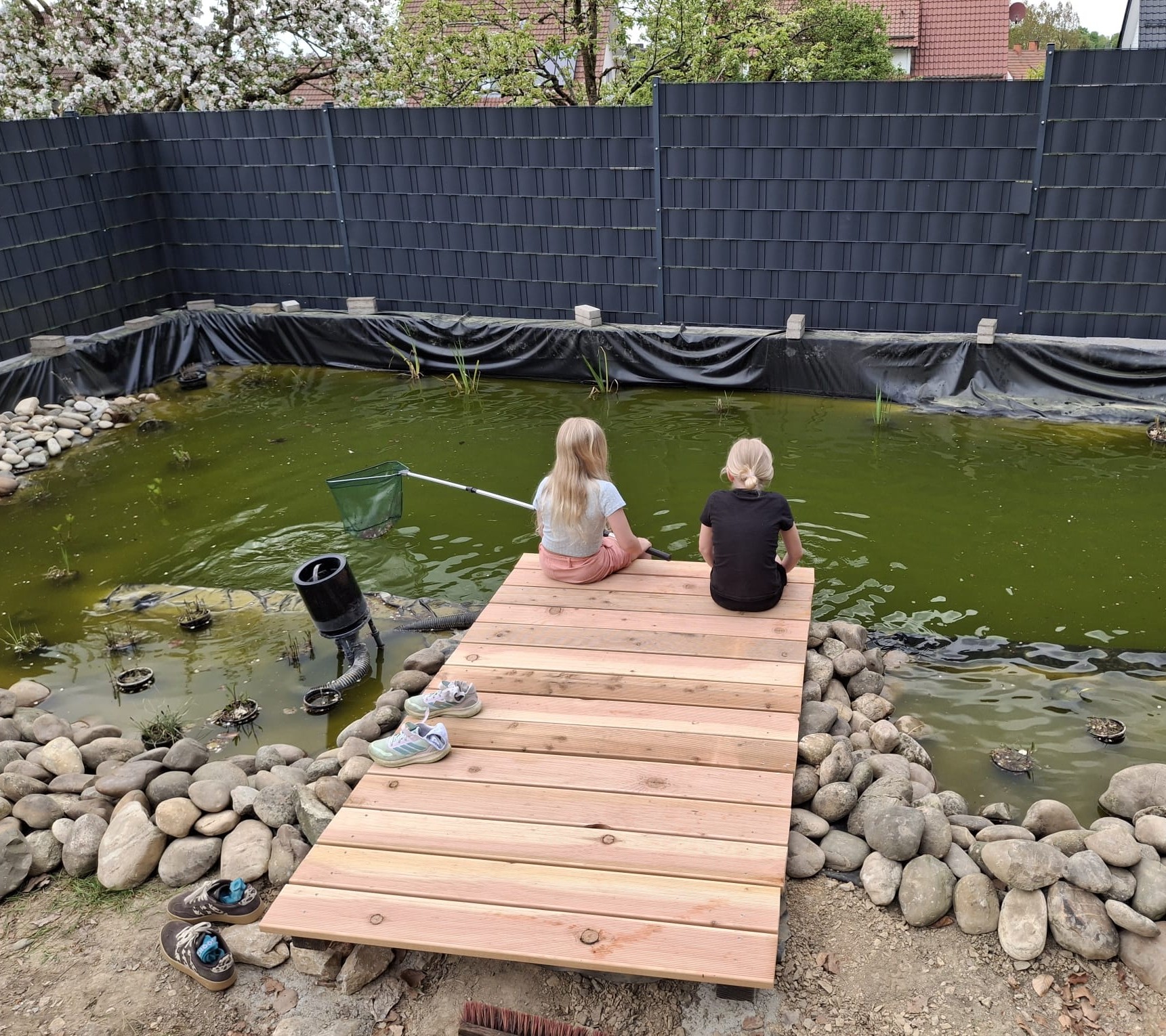 Zwei Kinder sitzen auf einem Holzsteg an einem kleinen Teich in einem eingezäunten Garten und halten ein Netz ins Wasser.