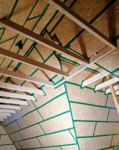 Dachboden mit sichtbaren Holzbalken und grün markierten Trennlinien auf Spanplatten.