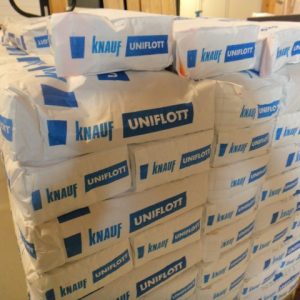 Mehrere gestapelte Packungen von Knauf Uniflott in einem Lagerraum, mit blauem Logo und Schriftzug auf weißer Verpackung, auf einer Holzpalette stehend.